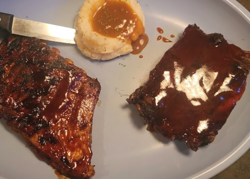 Two pieces of glazed ribs on a blue plate, accompanied by mashed potatoes and gravy. A knife rests beside the ribs.