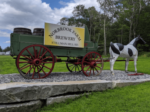 A vintage wagon with barrels and a sign for Norbrook Farm Brewery, next to a painted horse statue on a grassy area.