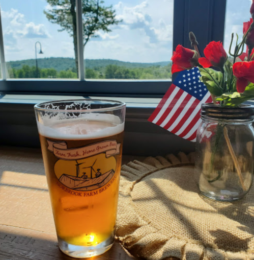 A glass of beer sits on a table with a view of mountains, alongside a small vase of flowers and a mini American flag.