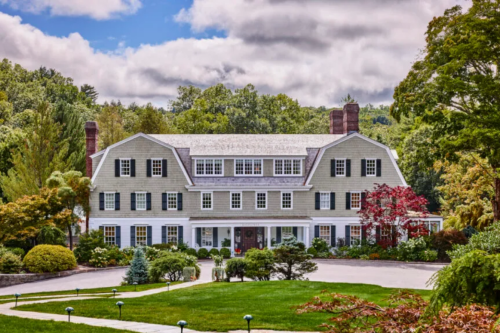 A large, elegant house with a green lawn, surrounded by trees and colorful shrubs under a partly cloudy sky.