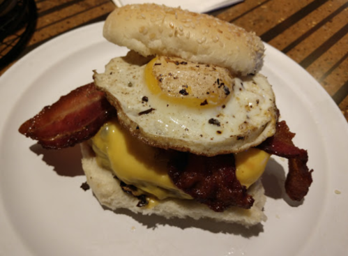 A close-up of a burger with a sesame seed bun, cheese, bacon, and a fried egg on top.