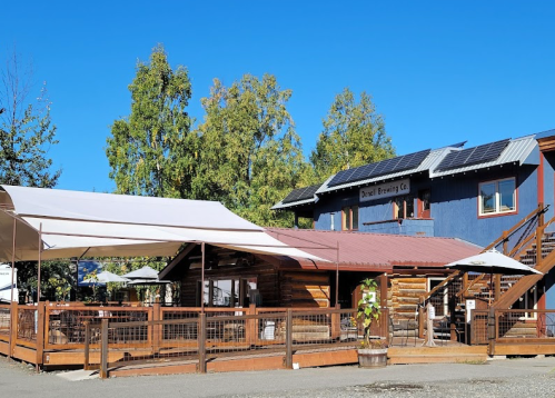 A rustic brewery with a wooden deck, solar panels, and outdoor seating under a clear blue sky.