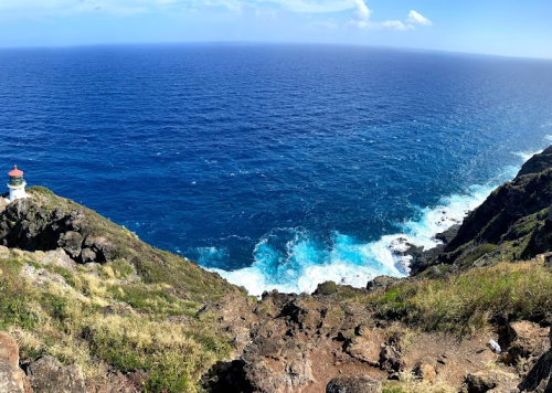 A panoramic view of a rocky coastline with a lighthouse overlooking the deep blue ocean and waves crashing against the shore.