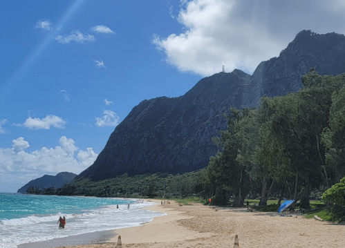 A scenic beach with turquoise water, sandy shore, and lush mountains under a partly cloudy sky. People enjoy the view.