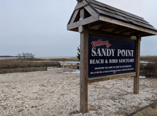 Sign for Sandy Point Beach & Bird Sanctuary, with a scenic view of the beach and marshland in the background.