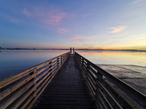 A wooden pier extends into a calm lake at sunset, with two figures standing at the end, surrounded by soft pastel skies.