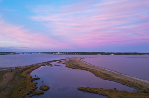 A serene landscape featuring a winding waterway, grassy banks, and a colorful sunset sky over a calm sea.