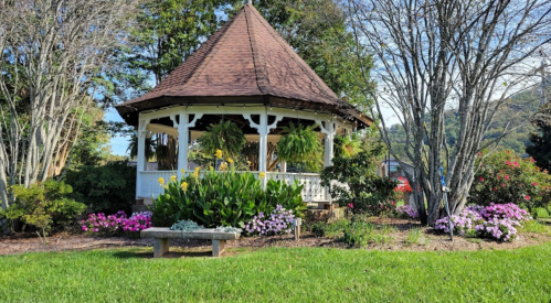 A charming gazebo surrounded by colorful flowers and greenery, set against a backdrop of trees and hills.