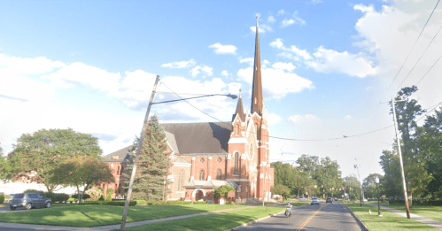 A tall church with a spire, surrounded by trees, on a sunny street with power lines and a few people walking.