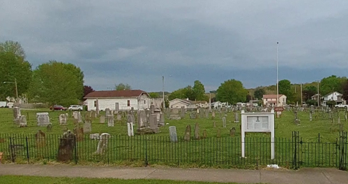 A cemetery with numerous gravestones, surrounded by a fence, and a few buildings in the background under a cloudy sky.