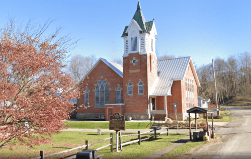 Historic brick church with a tall steeple, surrounded by trees and a grassy area, on a clear sunny day.