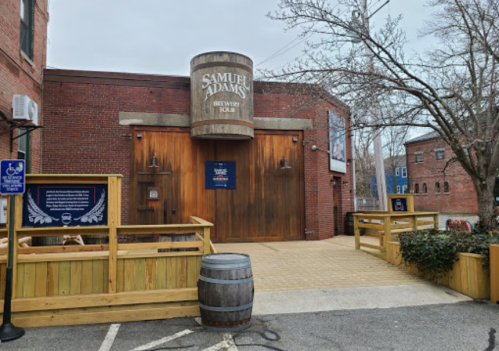 Exterior of the Samuel Adams Brewery, featuring a wooden entrance and a barrel, surrounded by a wooden fence.