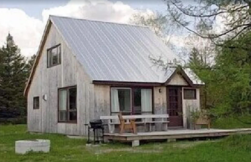 A rustic wooden house with a metal roof, surrounded by greenery and featuring a small porch with seating.