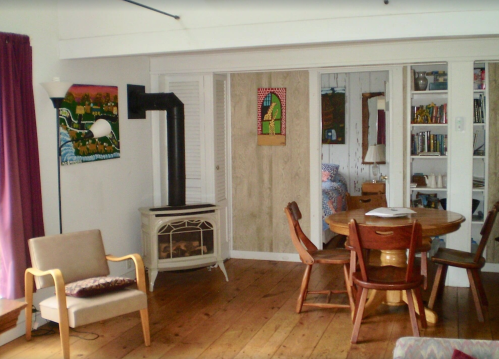 Cozy living space with a round wooden table, chairs, a lamp, and artwork on the walls, featuring wooden floors.