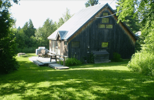 A rustic wooden cabin surrounded by greenery, featuring a deck and outdoor seating in a serene landscape.