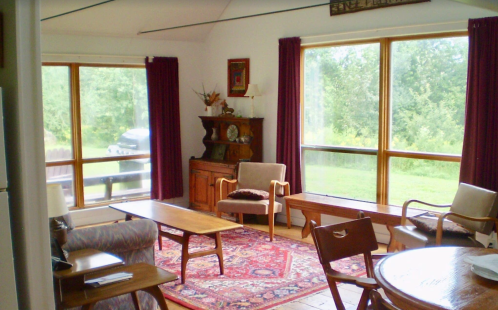 Cozy living room with large windows, wooden furniture, a rug, and a view of greenery outside.