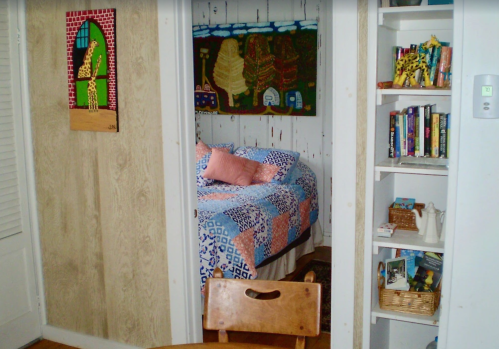 Cozy room with a bed, colorful quilt, bookshelf, and artwork on the walls, viewed from a small dining area.