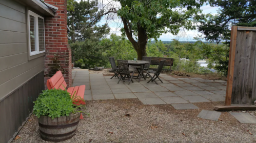 A patio with a wooden table and chairs, surrounded by greenery and a brick wall, featuring a red bench and gravel.