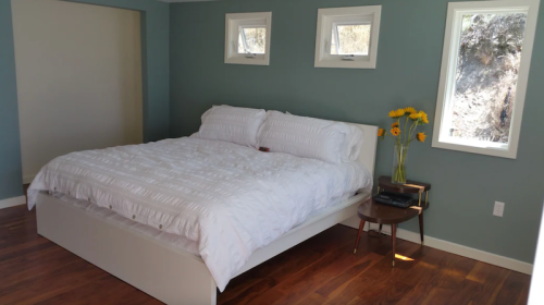 A bright bedroom with a large white bed, wooden floor, and a small table with sunflowers by the window.