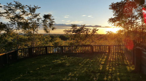 Sunset view from a wooden deck surrounded by trees, with golden light illuminating the landscape and clouds.