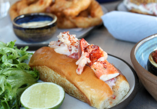 A lobster roll topped with lobster meat and seasoning, served with a lime wedge and a side of greens.