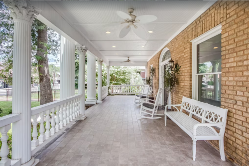 A spacious porch with white rocking chairs, a bench, and decorative columns, surrounded by greenery.