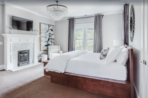 Bright bedroom featuring a large bed, fireplace, TV, and elegant chandelier, with neutral decor and large windows.