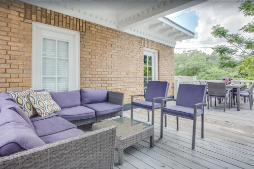A cozy outdoor seating area with a purple couch, chairs, and a glass table on a wooden deck surrounded by greenery.