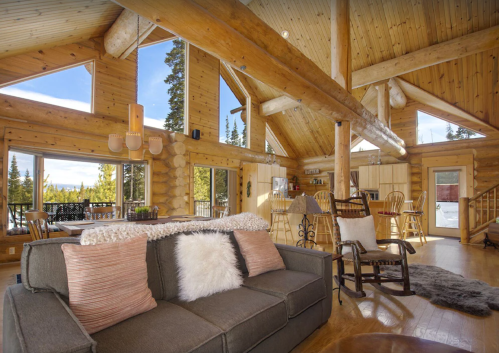 Cozy log cabin interior with wooden beams, large windows, and a comfortable sofa, surrounded by nature.