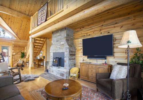 Cozy log cabin living room with wooden walls, a stone fireplace, a TV, and comfortable seating.