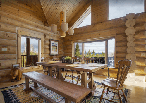 Cozy log cabin interior with a wooden dining table, chairs, and large windows showcasing a scenic view.