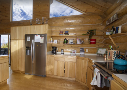 A modern kitchen with wooden walls, stainless steel fridge, open shelves, and a blue kettle on the stove.