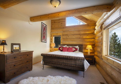 Cozy bedroom with log walls, a bed with decorative pillows, a dresser, and a window letting in natural light.