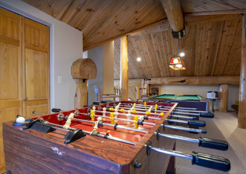 A cozy game room featuring a foosball table and a pool table, with wooden beams and a rustic ceiling.