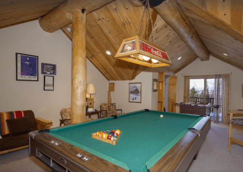 A cozy game room featuring a pool table, wooden beams, and comfortable seating with large windows.