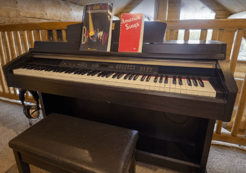 A digital piano with sheet music on top, a bench in front, and headphones resting beside it in a wooden room.
