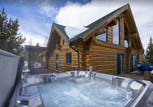 A log cabin with large windows and a hot tub in the foreground, surrounded by snow-covered trees and a blue sky.