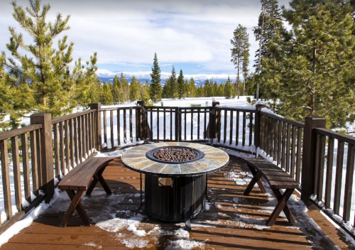 A snowy outdoor deck with a circular fire pit and wooden benches, surrounded by tall pine trees and mountains in the background.
