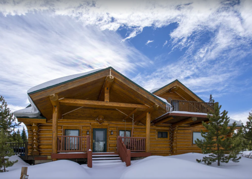 A log cabin with a large porch, surrounded by snow and pine trees, under a blue sky with wispy clouds.