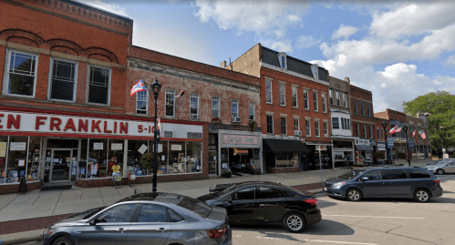 A street view of a small town with brick buildings, shops, and parked cars under a partly cloudy sky.