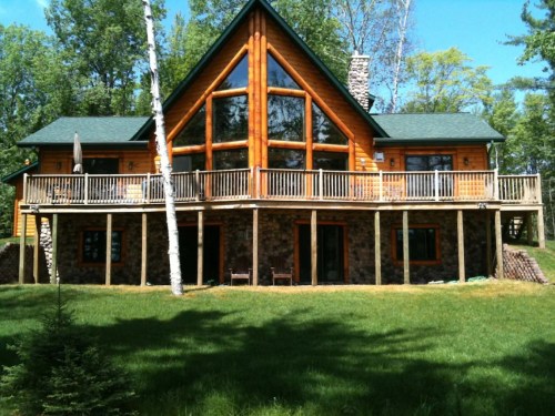 A large wooden cabin with a sloped roof, surrounded by trees and a grassy area, featuring a spacious deck.