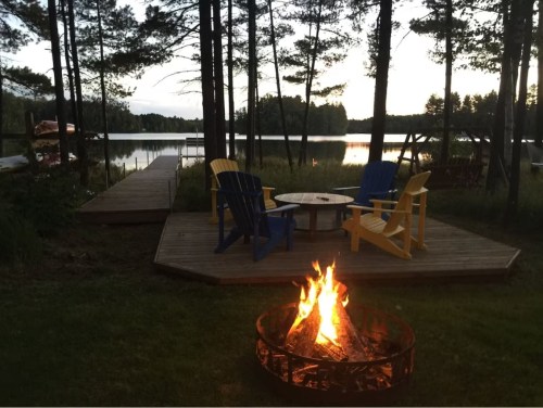 A cozy lakeside scene with a fire pit, colorful chairs, and a wooden dock surrounded by trees at sunset.