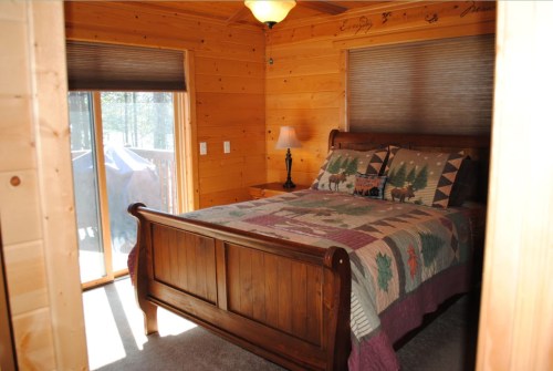 Cozy wooden bedroom with a quilted bedspread, lamp, and large windows overlooking a deck. Warm, rustic decor.