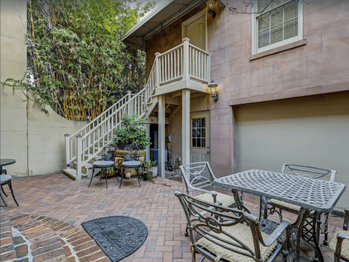 A cozy outdoor patio with a table and chairs, leading to a staircase and a garage, surrounded by greenery.