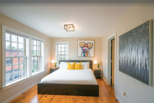 Bright bedroom with a large bed, yellow pillows, artwork on the wall, and windows letting in natural light.