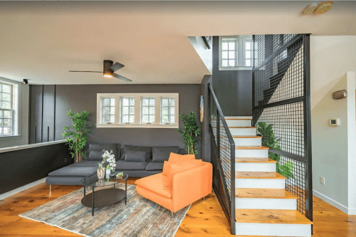 Modern living room with a gray sofa, orange chair, plants, and a staircase with a black railing. Bright and inviting space.