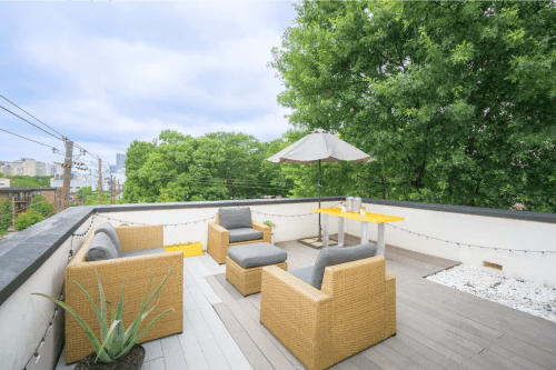 Rooftop patio with yellow table, chairs, and an umbrella, surrounded by greenery and city views.