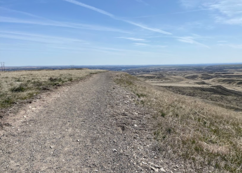 A gravel path winds along a grassy ridge, overlooking a vast, open landscape under a clear blue sky.