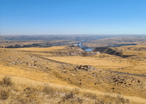 A panoramic view of a dry, golden landscape with rolling hills and a winding river in the distance under a clear blue sky.