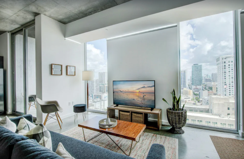 Modern living room with large windows, a TV, a coffee table, and a view of the city skyline. Bright and airy atmosphere.